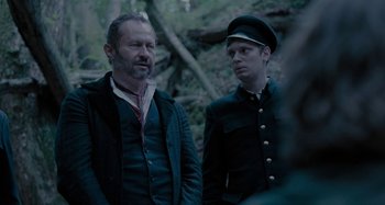 Movie still from “The Field Guide to Evil” (2018), directed by Can Evrenol – A man and a man in a uniform standing next to each other in the woods; Medium shot, Over the shoulder angle