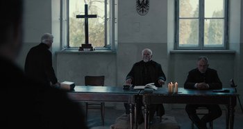 Movie still from “The Field Guide to Evil” (2018), directed by Can Evrenol – An old man sitting at a table in front of a crucifix; Wide shot, Over the shoulder angle