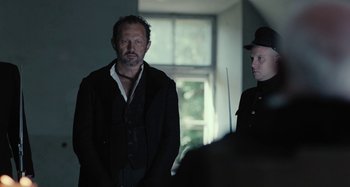 Movie still from “The Field Guide to Evil” (2018), directed by Can Evrenol – A man standing next to another man in a room; Medium shot, Over the shoulder angle