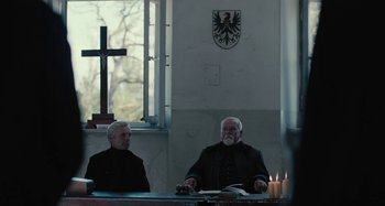Movie still from “The Field Guide to Evil” (2018), directed by Can Evrenol – Two men sitting at a table in front of a cross; Medium shot, Low angle