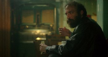 Movie still from “The Field Guide to Evil” (2018), directed by Can Evrenol – A man with a beard is holding a cigarette and a drink; Medium shot, Over the shoulder angle