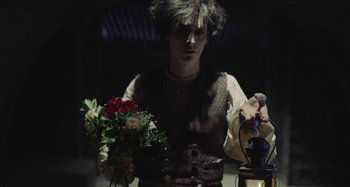 Movie still from “The Field Guide to Evil” (2018), directed by Can Evrenol – A man holding a bouquet of flowers in a dark room; Medium shot, Over the shoulder angle