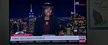 Movie still from “The First Purge” (2018), directed by Gerard McMurray – A woman sitting in front of a news desk in front of a city skyline at night; Medium shot, Over the shoulder angle