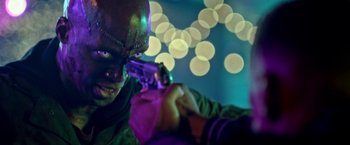 Movie still from “The First Purge” (2018), directed by Gerard McMurray – A man in a green mask holding a gun in front of lights; Close Up shot, Over the shoulder angle