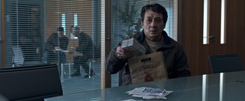 Movie still from “The Foreigner” (2017), directed by Martin Campbell – A man sitting at a table holding a bag of food; Medium shot, Over the shoulder angle