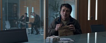Movie still from “The Foreigner” (2017), directed by Martin Campbell – A man sitting at a table holding a bag of money; Medium shot, Over the shoulder angle