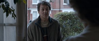 Movie still from “The Foreigner” (2017), directed by Martin Campbell – A person wearing a jacket near a building; Medium shot, Over the shoulder angle