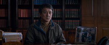 Movie still from “The Foreigner” (2017), directed by Martin Campbell – An older man sitting at a table in front of a book shelf; Medium shot, High angle