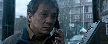 Movie still from “The Foreigner” (2017), directed by Martin Campbell – An older man talking on a cell phone while standing in front of a window; Close Up shot, Low angle