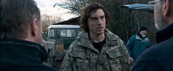 Movie still from “The Foreigner” (2017), directed by Martin Campbell – A man in camouflage jacket standing in front of a truck; Close Up shot, Over the shoulder angle