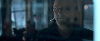 Movie still from “The Foreigner” (2017), directed by Martin Campbell – A man with a cigarette in his mouth looking out a window; Close Up shot, Over the shoulder angle
