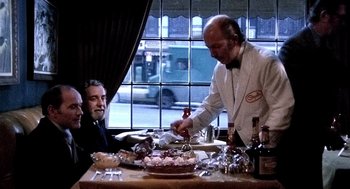 Movie still from “The French Connection” (1971), directed by William Friedkin – Two men sitting at a table with a cake on top of it; Medium shot, Over the shoulder angle