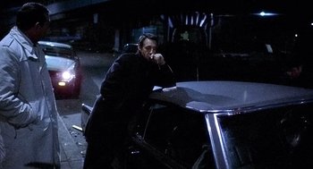 Movie still from “The French Connection” (1971), directed by William Friedkin – A man smoking a cigarette next to a parked car at night; Medium shot, Low angle