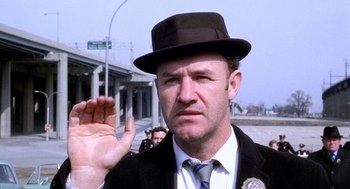 Movie still from “The French Connection” (1971), directed by William Friedkin – A man in a suit and a black hat is holding his hand up; Close Up shot, Over the shoulder angle