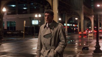 Movie still from “The Fugitive” (1993), directed by Andrew Davis – A man in a trench coat and tie standing on a sidewalk; Medium shot, Over the shoulder angle