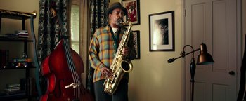 Movie still from “Popstar: Never Stop Never Stopping” (2016), directed by Akiva Schaffer – A man holding a saxophone while standing in a living room; Medium shot, Low angle