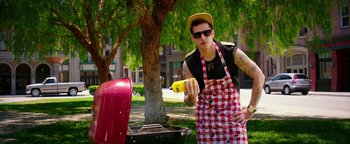 Movie still from “Popstar: Never Stop Never Stopping” (2016), directed by Akiva Schaffer – A man holding a banana while standing in front of a grill; Medium shot, High angle