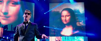 Movie still from “Popstar: Never Stop Never Stopping” (2016), directed by Akiva Schaffer – A man standing in front of a large painting of a woman; Medium shot, Over the shoulder angle