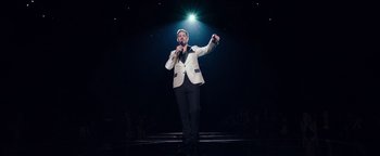 Movie still from “Popstar: Never Stop Never Stopping” (2016), directed by Akiva Schaffer – A man in a white jacket is holding a microphone; Wide shot, High angle