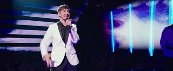 Movie still from “Popstar: Never Stop Never Stopping” (2016), directed by Akiva Schaffer – A man in a white jacket is singing into a microphone; Medium shot, Over the shoulder angle