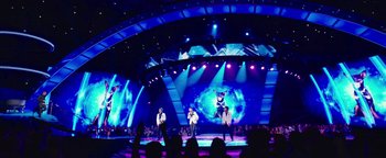 Movie still from “Popstar: Never Stop Never Stopping” (2016), directed by Akiva Schaffer – A group of people standing on a stage in front of a screen; Extreme Wide shot, Low angle