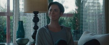 Movie still from “The Gift” (2015), directed by Joel Edgerton – A woman smiling for the camera in front of a lamp; Close Up shot, Over the shoulder angle