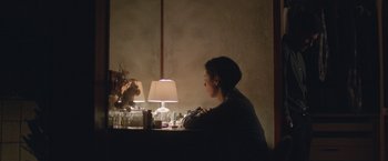 Movie still from “The Gift” (2015), directed by Joel Edgerton – A person sitting at a table with a lamp in a dark room; Medium shot, Over the shoulder angle
