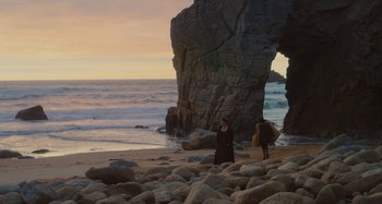 Movie still from “Portrait of a Lady on Fire” (2019), directed by Céline Sciamma – Two people walking on a beach near a rock arch; Extreme Wide shot, High angle