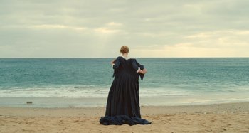 Movie still from “Portrait of a Lady on Fire” (2019), directed by Céline Sciamma – A woman in a black dress standing on the beach looking out at the ocean; Wide shot, High angle