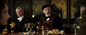 Movie still from “The Golden Compass” (2007), directed by Chris Weitz – An older man sitting at a table with food and wine; Medium shot, Over the shoulder angle
