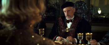 Movie still from “The Golden Compass” (2007), directed by Chris Weitz – An older man wearing a black hat sitting at a dinner table; Medium shot, Over the shoulder angle