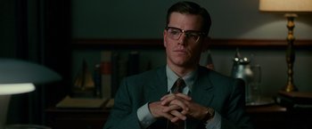 Movie still from “The Good Shepherd” (2006), directed by Robert De Niro – A man in a suit and tie sitting in front of an american flag; Close Up shot, Over the shoulder angle