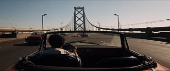 A man driving a car on a road with a bridge in the background