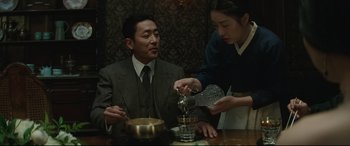 Movie still from “The Handmaiden” (2016), directed by Park Chan-wook – A man sitting at a table with a woman pouring something into a glass; Medium shot, Low angle