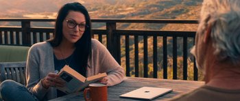 Movie still from “The Hero” (2017), directed by Brett Haley – A woman sitting at a table reading a book with an open laptop in front of her; Medium shot, High angle