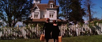 Movie still from “Practical Magic” (1998), directed by Griffin Dunne – Two women in black dresses holding suitcases in front of a white house; Wide shot, Low angle