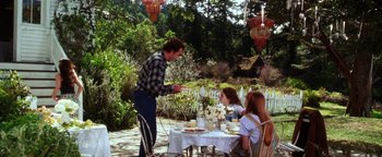 Movie still from “Practical Magic” (1998), directed by Griffin Dunne – A man standing next to a woman at an outdoor table; Wide shot, Over the shoulder angle