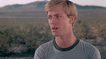 Movie still from “The Hills Have Eyes” (1977), directed by Wes Craven – A man with blonde hair wearing a gray shirt; Close Up shot, Low angle