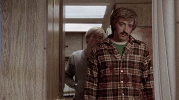 Movie still from “The Hills Have Eyes” (1977), directed by Wes Craven – A man in a plaid shirt standing next to another man in a green shirt; Medium shot, Over the shoulder angle