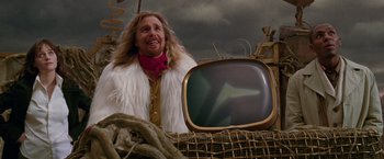 Movie still from “The Hitchhiker's Guide to the Galaxy” (2005), directed by Garth Jennings – A man in a white fur coat standing in front of an old tv; Medium shot, Low angle