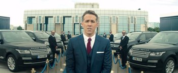 Movie still from “The Hitman's Bodyguard” (2017), directed by Patrick Hughes – A man wearing a suit and tie standing in front of a building; Medium shot, Over the shoulder angle