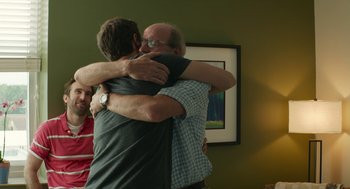 Movie still from “The Hollars” (2016), directed by John Krasinski – A couple of men hugging each other in a living room; Medium shot, Over the shoulder angle