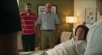 Movie still from “The Hollars” (2016), directed by John Krasinski – Two men standing in front of a woman in a hospital bed; Medium shot, Over the shoulder angle