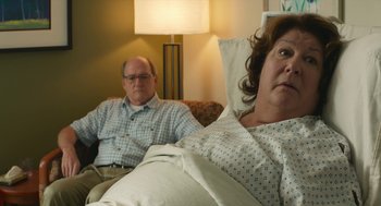 Movie still from “The Hollars” (2016), directed by John Krasinski – An older man sitting next to an older woman in a hospital bed; Medium shot, Over the shoulder angle