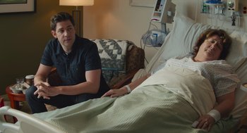 Movie still from “The Hollars” (2016), directed by John Krasinski – A man sitting next to a woman in a hospital bed; Medium shot, Over the shoulder angle