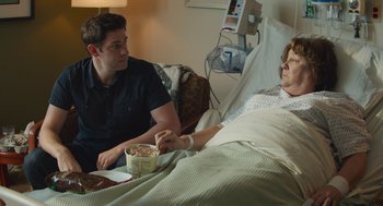 Movie still from “The Hollars” (2016), directed by John Krasinski – A man sitting next to an older woman in a hospital bed; Medium shot, Over the shoulder angle