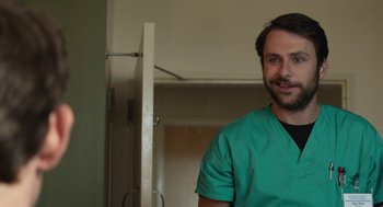 Movie still from “The Hollars” (2016), directed by John Krasinski – A man wearing a green scrub suit standing in a room; Close Up shot, Over the shoulder angle