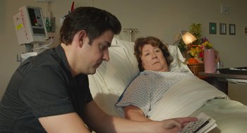 Movie still from “The Hollars” (2016), directed by John Krasinski – A man and a woman in a hospital bed looking at a computer; Medium shot, Over the shoulder angle
