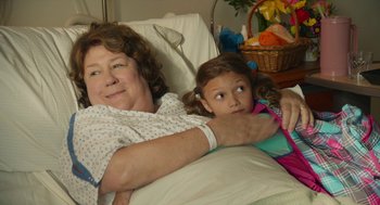 Movie still from “The Hollars” (2016), directed by John Krasinski – An older woman laying in a bed with a young girl; Medium shot, Over the shoulder angle