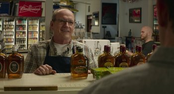 Movie still from “The Hollars” (2016), directed by John Krasinski – An older man sitting at a table in front of several bottles of alcohol; Medium shot, Over the shoulder angle